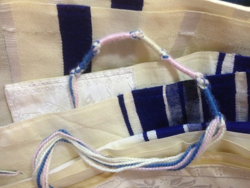 A folded tallit (prayer shawl) with blue and white stripes, with one corner showing its tzitzit (fringes), tied in five equal sections of blue, pink, white, pink, and blue.